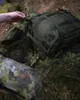 A person's hand is adjusting a buckle on a green backpack lying on the forest floor. Produktbild-Vorschau