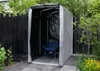 A garden shed with a wheelbarrow inside, set in a garden with a wooden fence and plants. Produktbild-Vorschau