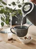 Hot water being poured from a kettle into a mug with a tea bag, next to a vase of eucalyptus leaves and sugar cubes. Produktbild-Vorschau
