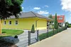 A bright yellow house with a gray roof is bordered by a dark gray metal fence. A paved driveway runs alongside the fence, leading towards another house with a red roof and solar panels. The scene is set on a sunny day with a blue sky and scattered clouds. Produktbild-Vorschau