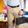 A man in a light blue shirt and tan pants stands in a room with a blurred background of balloons and furniture, holding a black gift bag. Anteprima dell’immagine prodotto