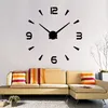 A large modern wall clock is displayed on a white wall above a sectional sofa with decorative pillows. Produktbild-Vorschau