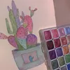 A watercolor painting of a cactus in a pot, next to a palette of shimmering watercolor paints. Voir les images du produit