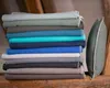 A stack of folded blankets in various shades of blue, green, and gray, with a green pillow standing to the right. Produktbild-Vorschau