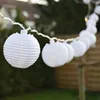 A string of white paper lanterns lit with warm white LEDs hangs outdoors, strung between two poles. The background shows greenery and a wooden fence. Produktbild-Vorschau