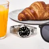 A watch, croissant, and orange juice are laid out on a white tablecloth, suggesting a breakfast setting. Produktbild-Vorschau