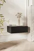 A black floating nightstand with a fluted front is mounted on a light-colored wall. It holds two books and a vase with dried flowers. A plant is visible on the left, and a portion of a bed is on the right. The floor is made of light wood planks. Produktbild-Vorschau