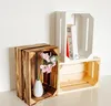 Three wooden crates are arranged against a white background. One crate on the left is dark brown and contains a white vase with pink flowers. Another crate is white and sits on top of a lighter brown crate. Produktbild-Vorschau