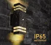 A black outdoor wall light with gold accents is mounted on a dark stone wall during a rain shower. Produktbild-Vorschau