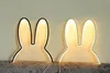 Two bunny ear-shaped night lights are displayed on a surface against a wall. Produktbild-Vorschau