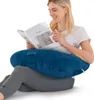 A woman is sitting on the floor, reading a book while using a U-shaped pillow for support. Produktbild-Vorschau