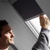 A man is adjusting a dark blue blackout roller blind on a skylight window. Produktbild-Vorschau