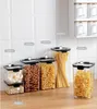 A collection of clear plastic food storage containers filled with various dry goods, arranged on a countertop against a tiled backsplash. Produktbild-Vorschau