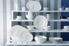 A collection of white ceramic dishes and cups displayed on white shelves against a striped blue and white background. Produktbild-Vorschau
