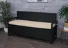 A black poly-rattan storage bench with a beige cushion is placed on a wooden floor in front of a grey wooden wall. There are plants on either side of the bench. Podgląd zdjęcia produktu