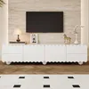 A white TV stand with drawers and doors, featuring a scalloped design, is shown in a living room setting with a TV mounted on a textured wall above it. The floor is wooden, and a rug with a black and white pattern is in the foreground. Produktbild-Vorschau