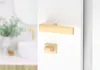 A close-up view of a gold-colored door handle and lock mechanism mounted on a white door. In the blurred background, a bathroom sink and faucet are visible. Produktbild-Vorschau
