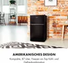 A black refrigerator is shown in a room with a brick wall and a patterned rug. Produktbild-Vorschau
