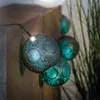 A string of solar-powered decorative lights with spherical, perforated shades in a teal and silver color scheme, hanging near a stone wall and tall grass. Produktbild-Vorschau