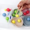 A hand using chopsticks to pick up a yellow ball from a beige heart-shaped bowl, surrounded by other colorful balls and bowls. Produktbild-Vorschau