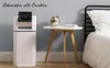 A white dehumidifier with a black fan is placed next to a bedside table with a book and a small plant on it, in a bedroom setting. Voir les images du produit