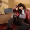 A woman poses next to a Marshall speaker in a dimly lit room with warm lighting. Produktbild-Vorschau