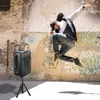 A man is jumping in the air in front of a graffiti-covered wall, with a speaker on a tripod to his left. Produktbild-Vorschau