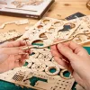 Hands assembling a wooden model kit, likely a vehicle, with various wooden parts and gears spread out on a table. Podgląd zdjęcia produktu