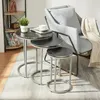 A set of three nesting tables with a marble-like black top and silver metal legs are placed on a rug next to a gray armchair. A plant and books are on a shelf in the background. Anteprima dell’immagine prodotto