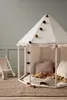 A children's play tent with decorative pom-poms, set up on a rug with a book and blanket inside. A toy stroller is visible in the background. Produktbild-Vorschau