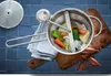 A top-down view of a food mill with vegetables inside, placed on a wooden cutting board on a blue tiled surface. Produktbild-Vorschau