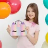 A young girl is holding a unicorn-themed handbag in front of a white background with colorful balloons. Voir les images du produit