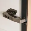 A close-up view of a cabinet hinge attached to a white door and a black frame. Produktbild-Vorschau