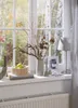 A windowsill decorated with autumn-themed items, including a vase with dried flowers, a bowl of apples, books, candles, a framed picture, and a lamp, with a view of trees outside. Podgląd zdjęcia produktu