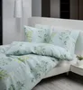 A neatly made bed with a floral pattern duvet and pillows, next to a floor lamp and a nightstand with a small plant. Produktbild-Vorschau