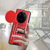 A hand holding a red phone case with a camera lens cover and a ring holder, with a blurred background of an indoor setting. Produktbild-Vorschau