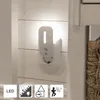 A white LED night light with a motion sensor plugged into a wall outlet, mounted on a white paneled wall. Below the light are four icons representing features: LED, sensor, home with an arrow, and a person with motion waves. Podgląd zdjęcia produktu