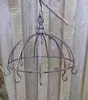 A rustic metal hanging basket with hooks, suspended from a branch against a wooden fence. Produktbild-Vorschau