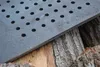 A close-up view of a perforated cast iron grate resting on rough-hewn logs. Náhled obrázku produktu