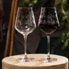 Two wine glasses, one empty and one filled with red wine, are placed on a wooden surface with a blurred background of greenery and wooden poles. Produktbild-Vorschau