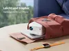 A white projector is placed in an open brown bag, with a book underneath it. A smartwatch with an orange strap is in the foreground. Produktbild-Vorschau