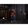 A couple stands outside a modern wooden house at night, illuminated by outdoor lighting. Produktbild-Vorschau