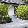 A paved walkway leads to a house with a garage, bordered by small fences and greenery. A child's bicycle is parked near the house. Náhled obrázku produktu