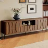 A wooden TV cabinet with decorative front panels and black metal legs, placed in a living room setting with artwork on the wall and a vase with flowers on top. Anteprima dell’immagine prodotto