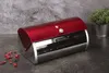 A red and silver bread box is displayed on a dark countertop with a gray textured background. A small potted plant is visible in the upper right corner. Náhled obrázku produktu