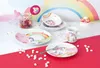 A collection of unicorn-themed tableware, including plates, bowls, and a mug, arranged on a table with marshmallows scattered around. A rainbow backdrop is visible. Anteprima dell’immagine prodotto