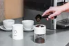A hand is pouring coffee beans into a coffee grinder, with two cups and saucers in the background. Podgląd zdjęcia produktu