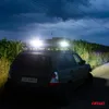 A dark, moody shot of a green SUV with bright auxiliary lights on its roof rack, parked on a dirt road next to a cornfield. Anteprima dell’immagine prodotto