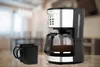 A coffee maker and two mugs on a wooden table, with a blurred kitchen background. Produktbild-Vorschau