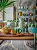 A collection of ceramic tableware, including mugs, a pitcher, and bowls, arranged on a wooden table in front of a stone wall. Produktbild-Vorschau